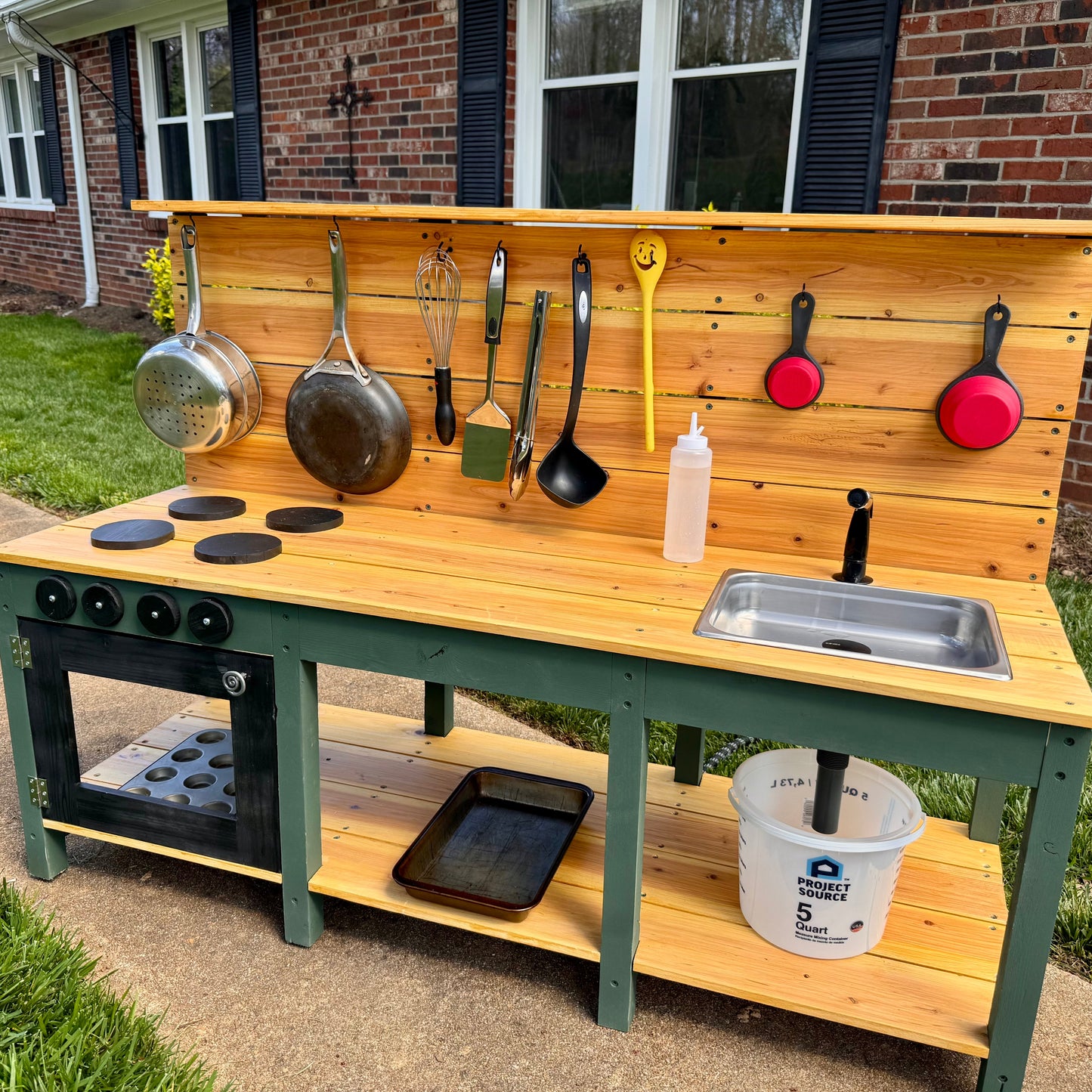Wooden play kitchen set with pots, pans, and utensils on a patio.
